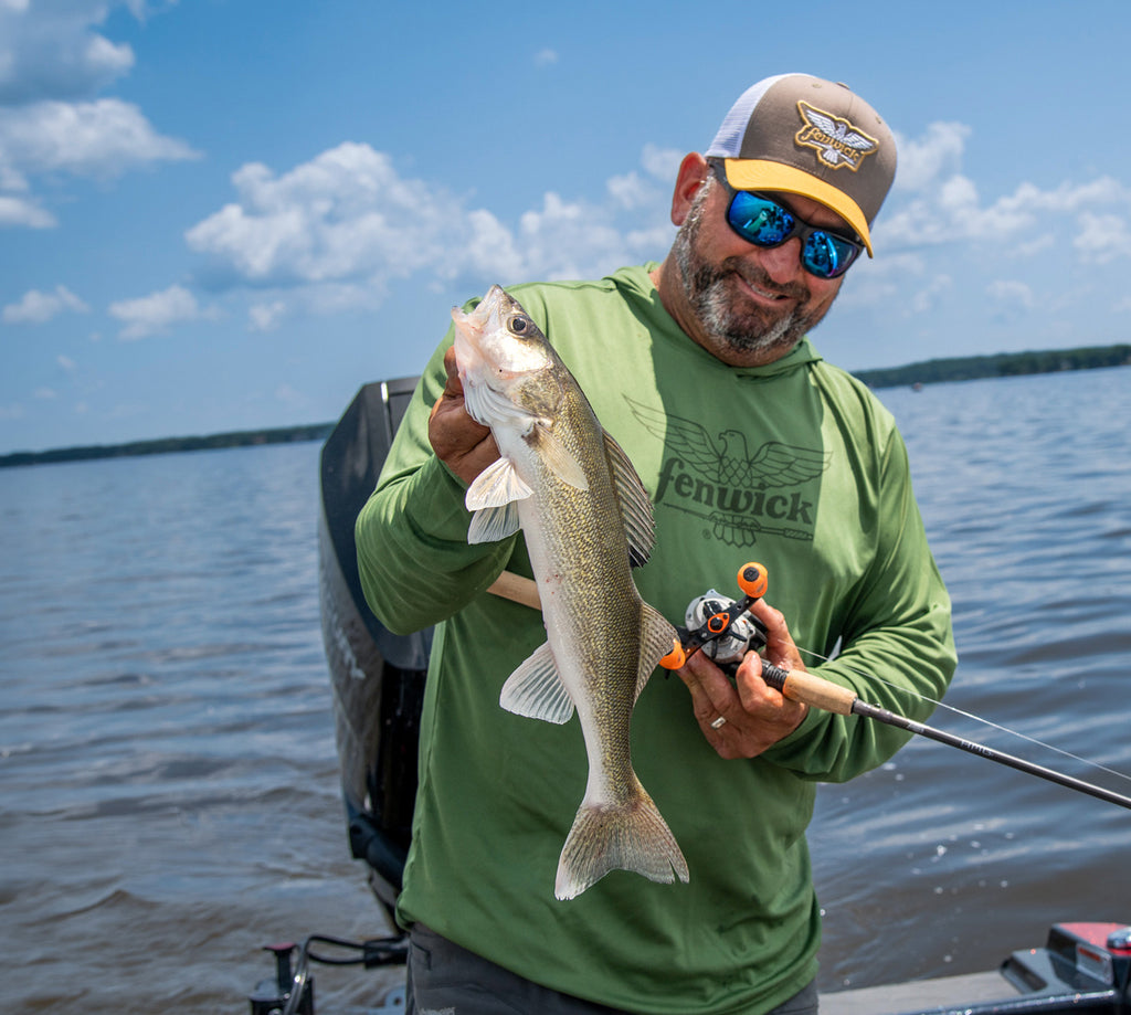 Man holding a walleye on a boat with a lake and sky in the background wearing Fenwick branded apparel