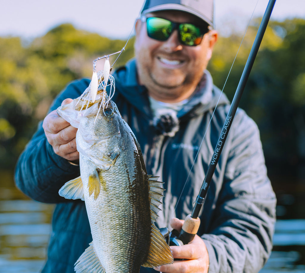 Man holding a large fish by a lake with trees in the background and a Fenwick World Class Rod. 