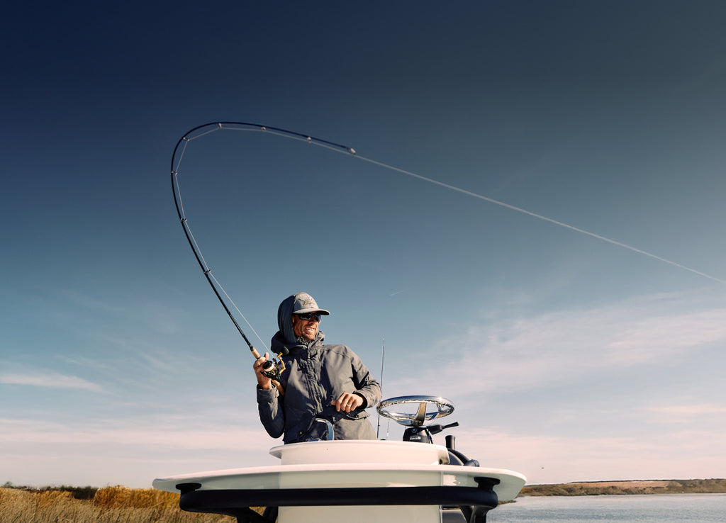 Person fishing on a boat with a curved fishing rod against a clear sky.